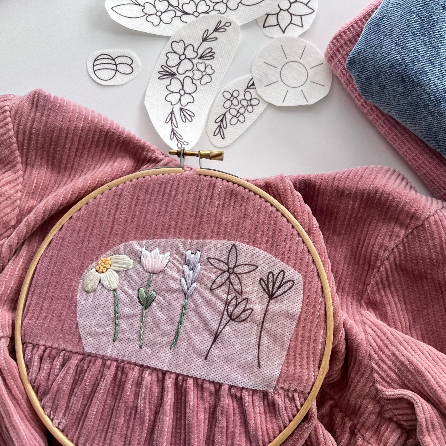 Set of 12 embroidery patches for upcycling clothes are scattered in the background, a floral design is being stitched onto a pink children's dress