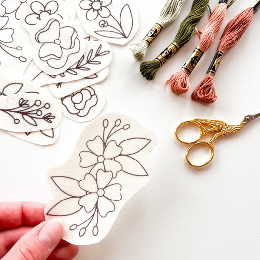 a single floral stick and stitch design consisting of two full flowers, leaves and buds is being held in the foreground over a white background, the other designs, gold stork scissors and 4 DMC pink and green threads are in the background